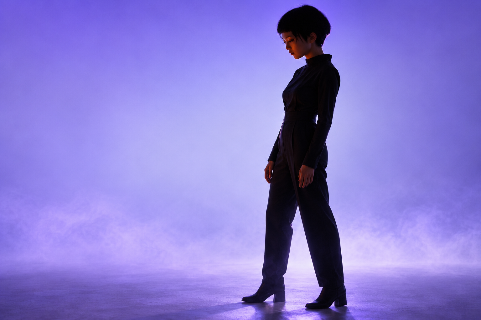 A woman with a black bob in dark tailoring, standing in lilac studio haze